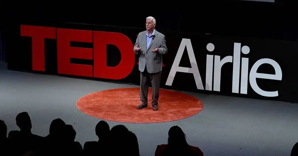 Neil Taft speaking on the TEDxAirlie stage in front of a large TEDxAirlie sign and audience.